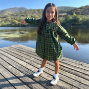 Young girl in a green checkered dress standing on a wooden dock by a lake.